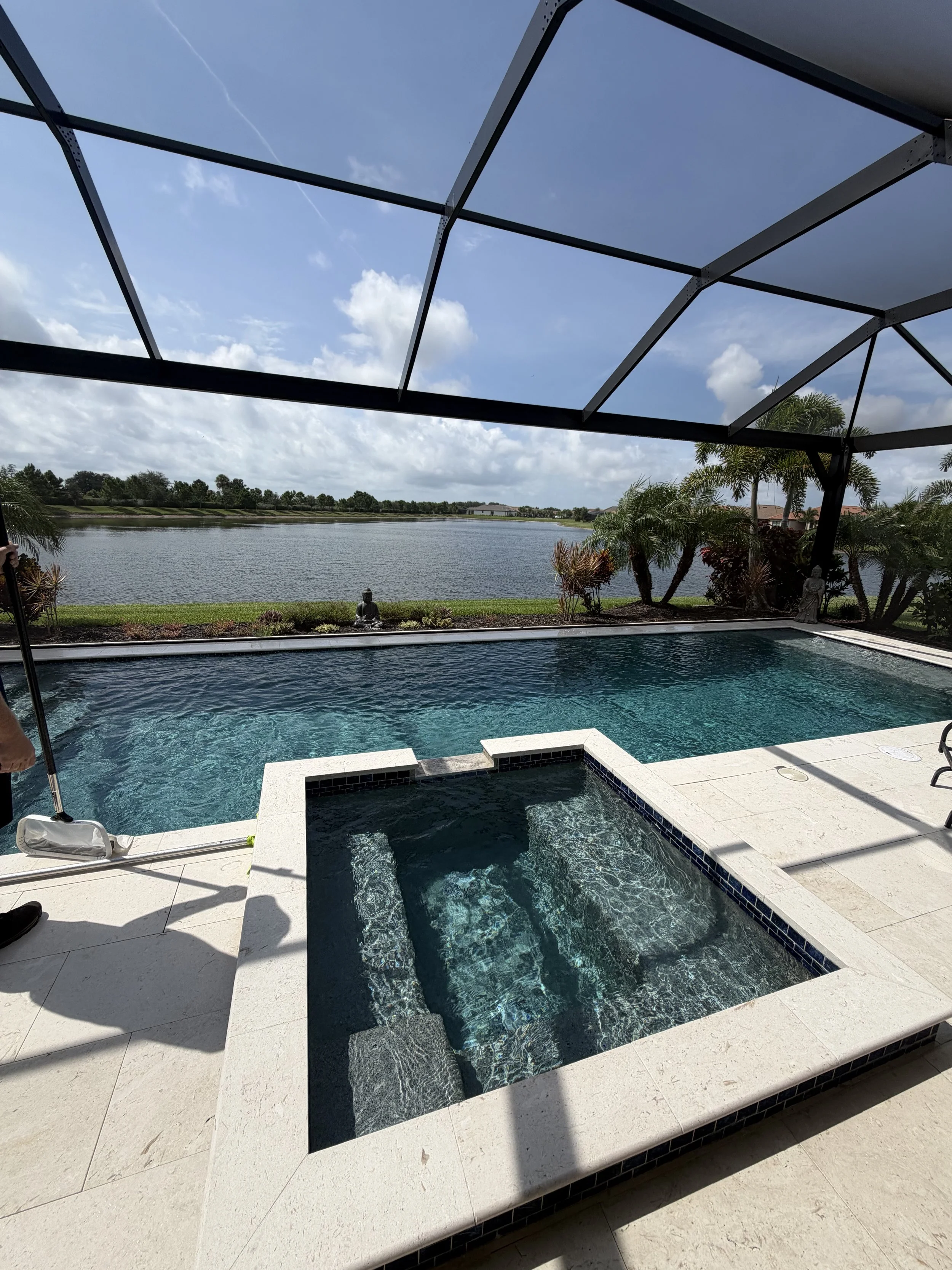 A backyard swimming pool with green water, surrounded by a textured concrete deck. A woman is standing nearby with a net on a pole, and there are lush trees and bushes in the background.