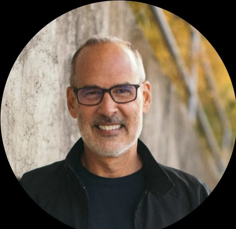 A smiling middle-aged man with glasses and a beard, standing outdoors against a brick wall with autumn trees in the background.