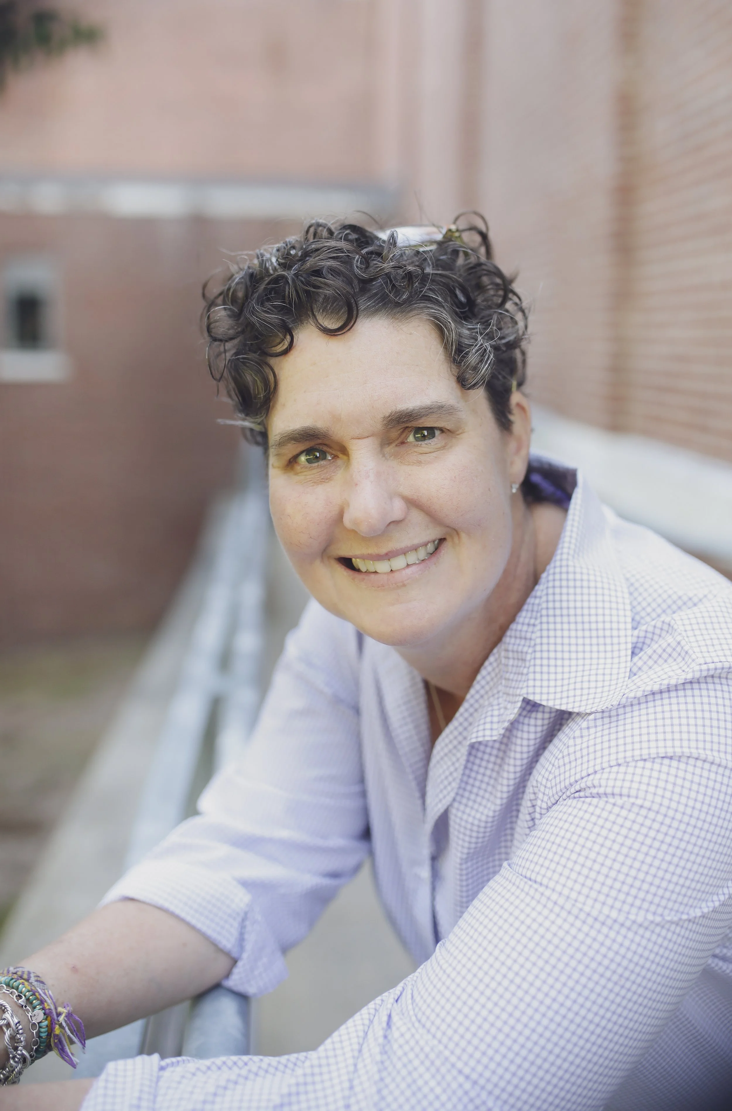 A woman with short, curly dark hair and light skin smiling at the camera. She is wearing a light-colored checkered shirt and is sitting on a metal railing outdoors, with a brick building in the background.