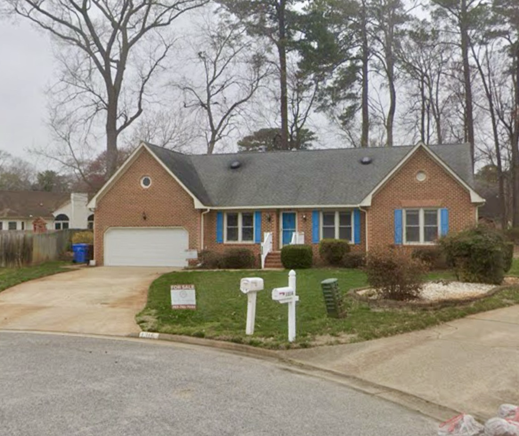 A brick house with a dark shingle roof, blue window shutters, a white front door with a small porch, and a two-car garage. The front yard has lawns, bushes, trees, and a sidewalk with a mailbox and a few signs.