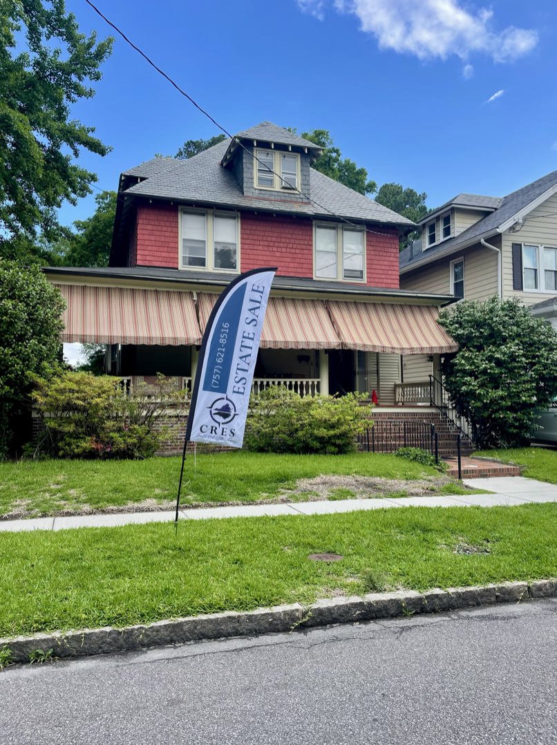 A large house with a red-tiled main section and an attic with dormer windows, a striped awning covering the porch, and a sign indicating a real estate sale.