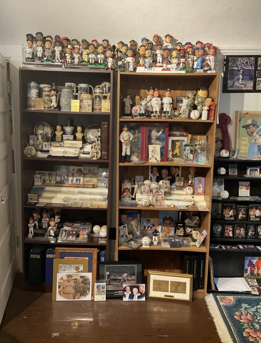A large collection of baseball memorabilia, including bobblehead dolls on top of two shelves, baseball cards, figurines, and framed photos displayed in a room.