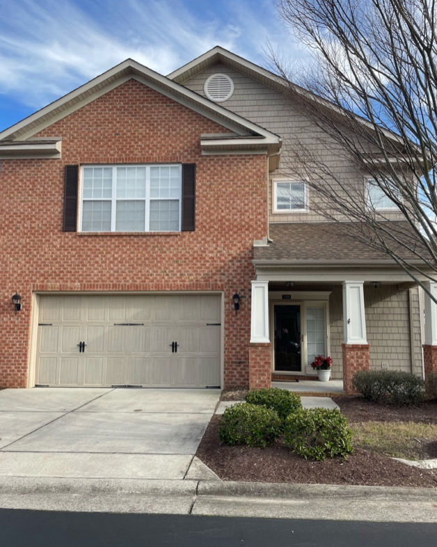 A two-story house with a brick facade on the left and beige siding on the right, featuring a closed garage door, porch with white columns, and potted plants at the entrance, under a partly cloudy sky.