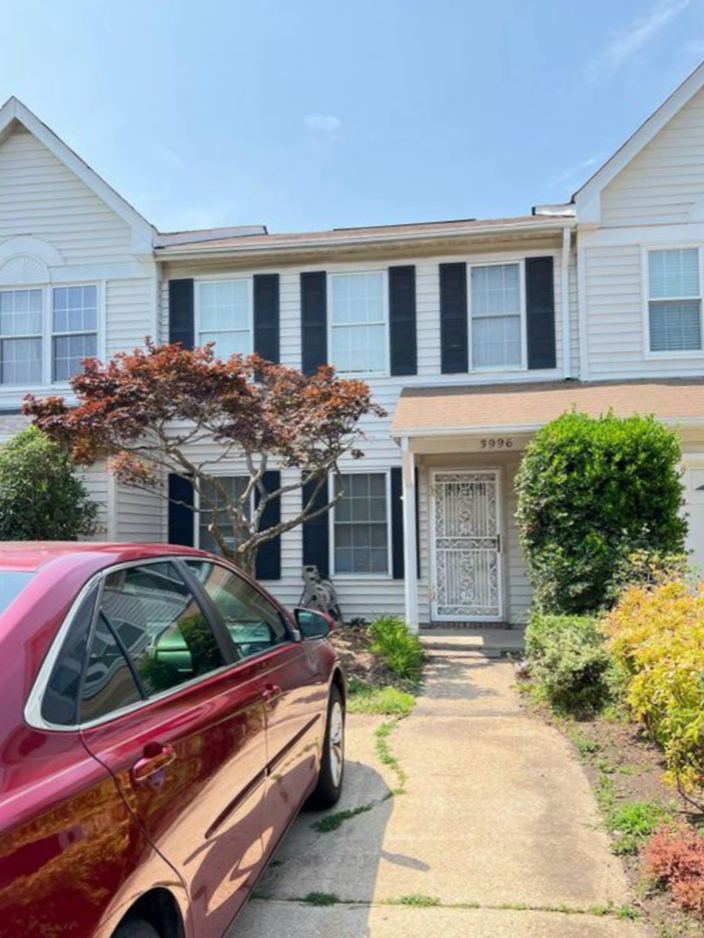 A red car parked in front of a two-story white house with black shutters, a small front yard with bushes and a tree, and a concrete walkway leading to the front door with a decorative metal security door.
