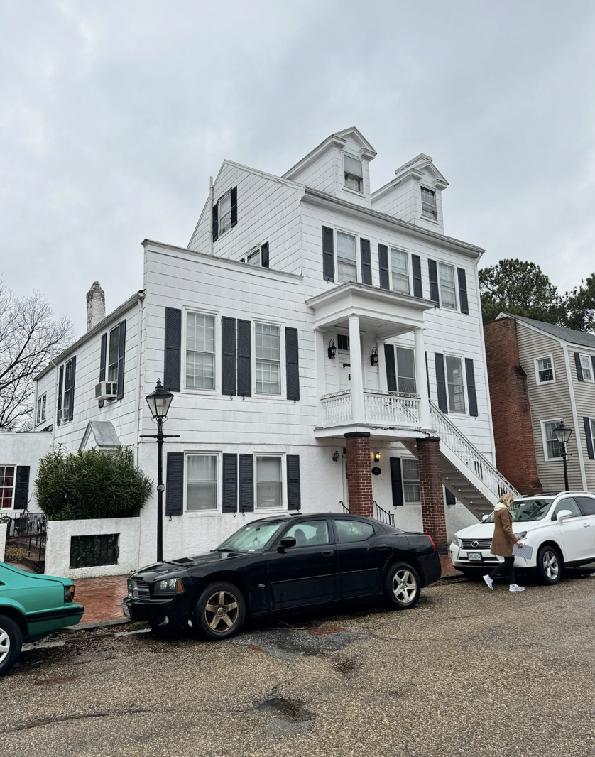 A white multi-story house with black shutters, a brick column porch, and a staircase leading to the second-floor entrance. Several cars are parked in front, and a woman is walking near a white SUV on a cloudy day.