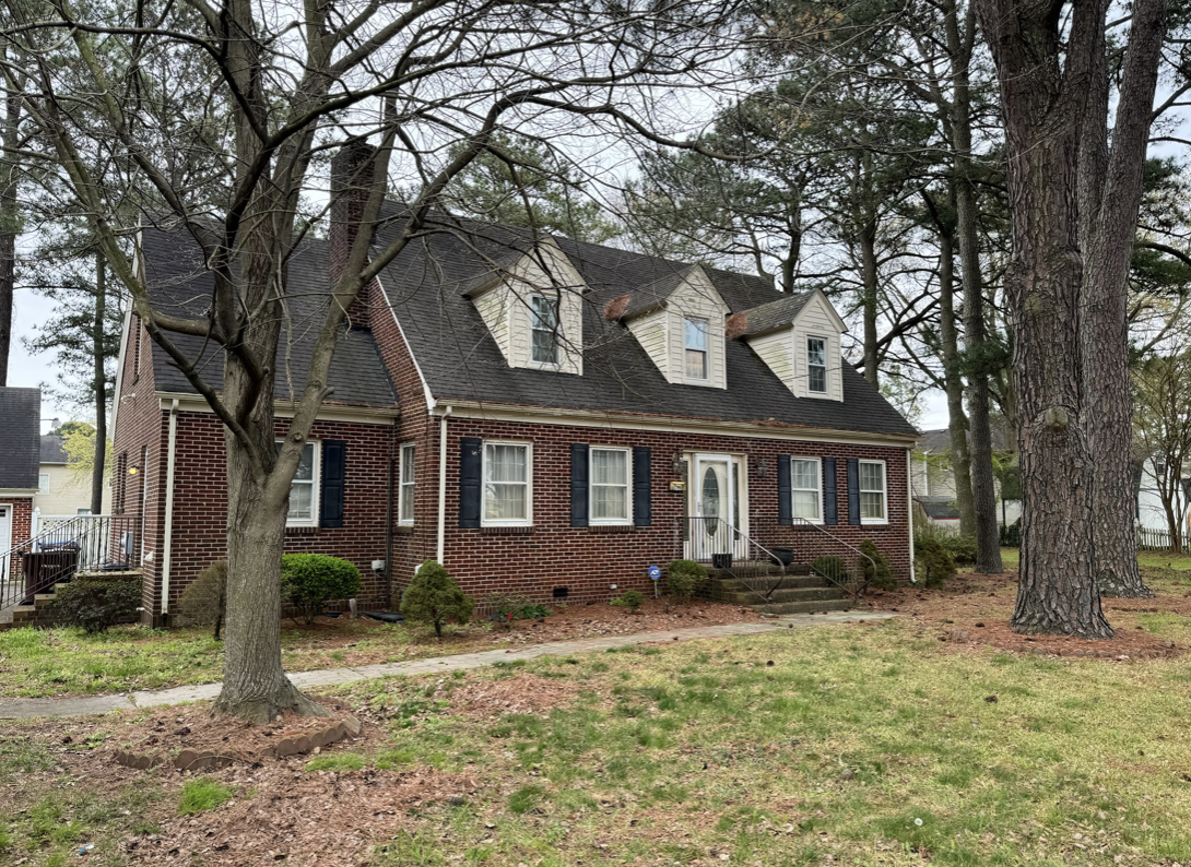 A brick house with three dormer windows on the roof, surrounded by trees and a well-maintained yard with small bushes, a sidewalk, and steps leading to the front door.