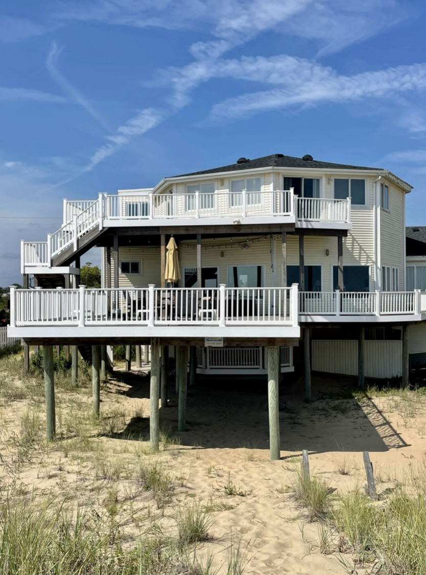 A white beach house raised on stilts with multiple decks and stairs, situated on sandy terrain with some grass and clear sky in the background.