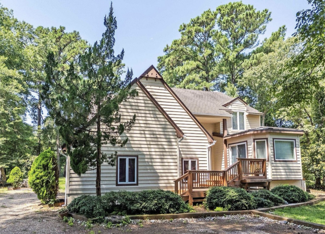 A beige house with a small wooden deck, surrounded by green trees and bushes, under a clear blue sky.
