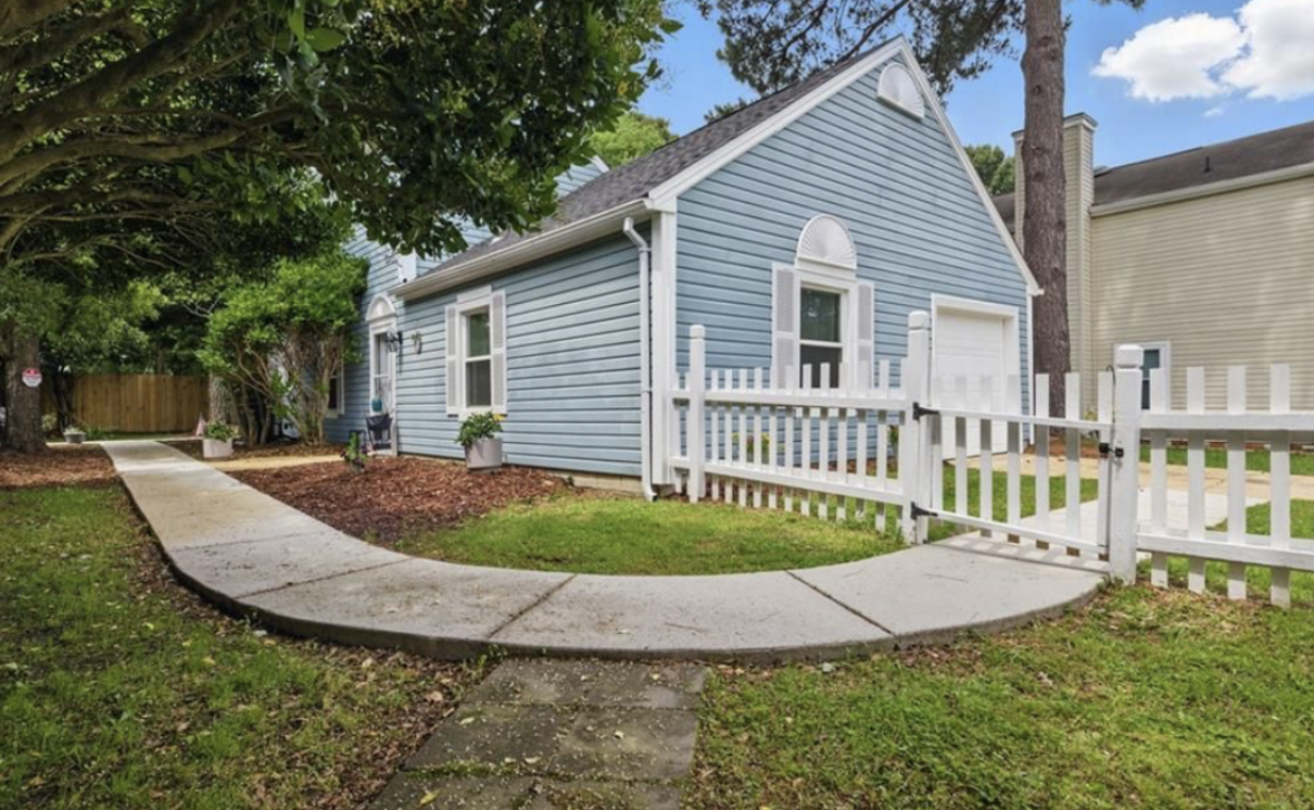 A blue house with white trim, a white picket fence gate, a concrete sidewalk, and surrounding green grass and trees.