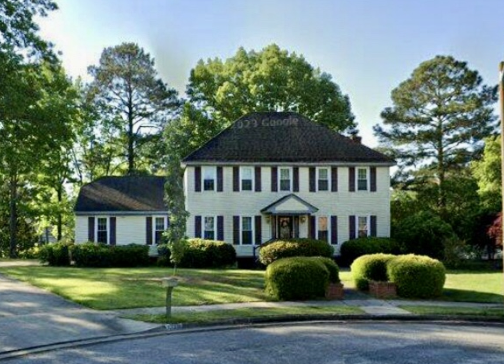 Bright two-story white house with dark shutters, surrounded by green trees and bushes, on a sunny day.