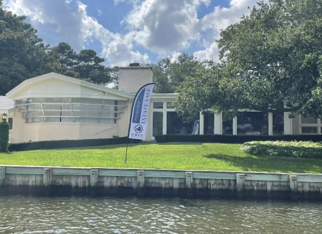 Modern house with large windows, lawn, and a 'For Sale' flag along a waterway with a dock.