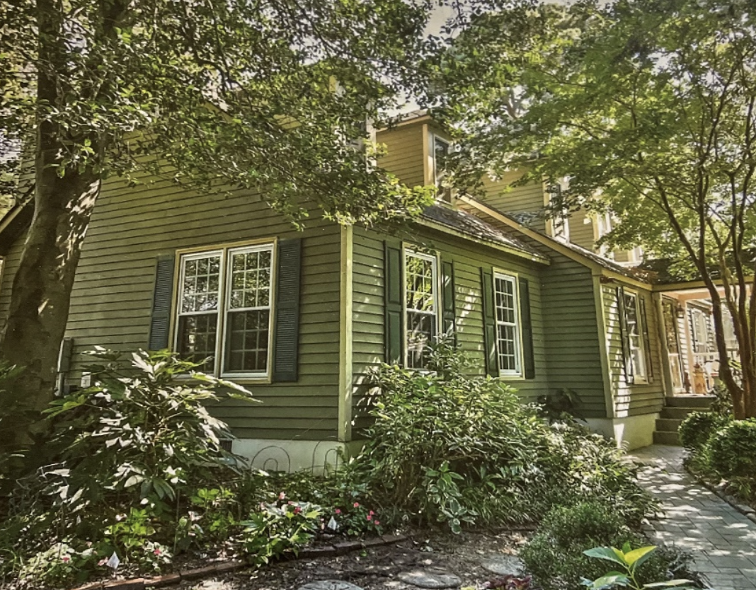 A green two-story house with black shutters, surrounded by trees and shrubs, with a stone pathway leading to the entrance.