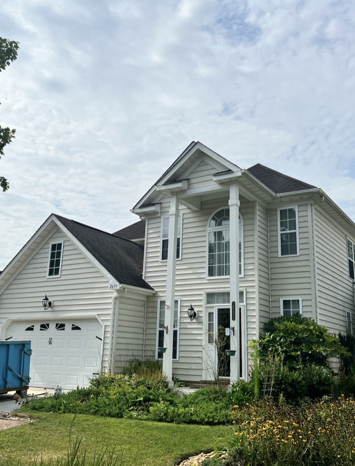 A tall, white, two-story house with a gabled roof, front porch columns, and large windows, surrounded by greenery and a well-maintained lawn.