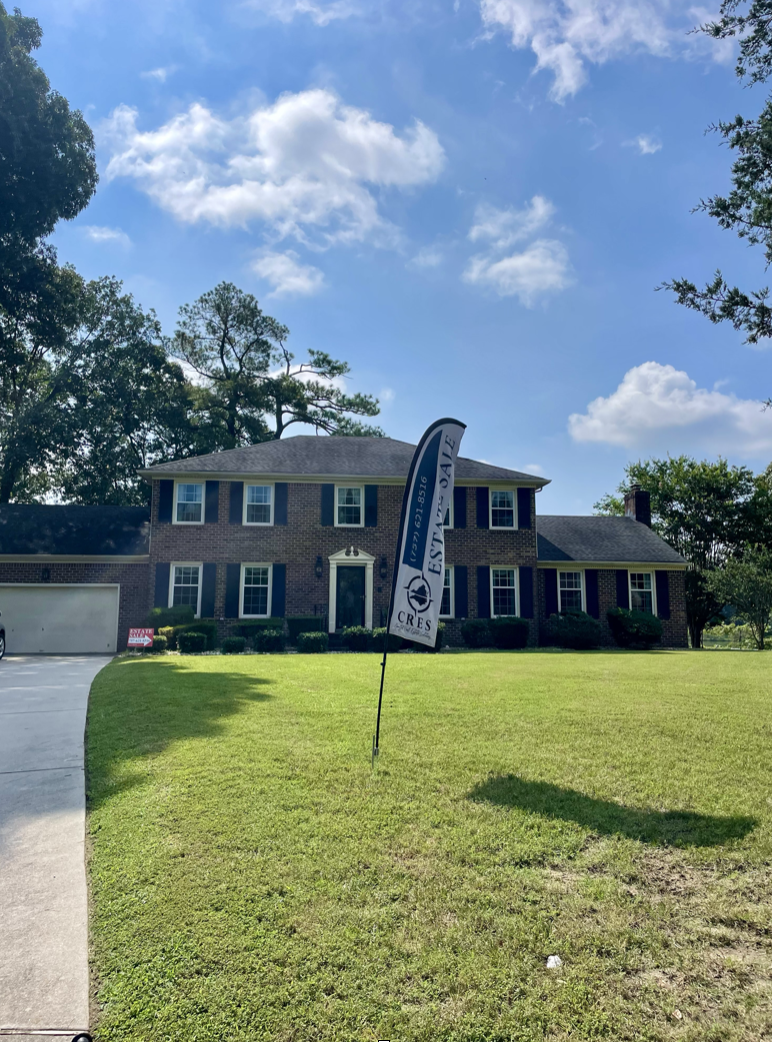 Two-story brick house with white trim, surrounded by a well-maintained lawn, with a black and white real estate sign in front reading 'For Sale,' under a partly cloudy blue sky with trees in the background.