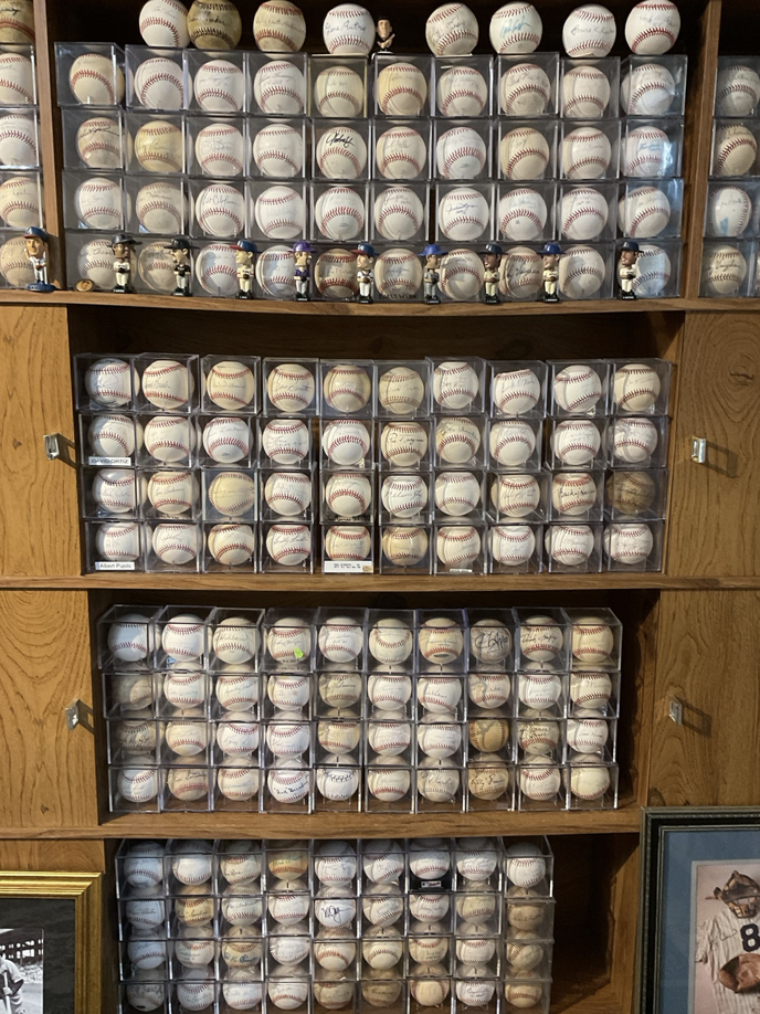 A wooden display cabinet with multiple shelves holding dozens of baseballs in clear plastic cases, some signed or marked with signatures or inscriptions.