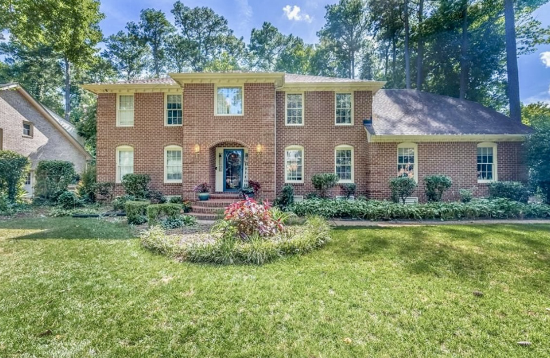 Front view of a two-story brick house with a well-maintained lawn and garden, surrounded by tall trees under a blue sky