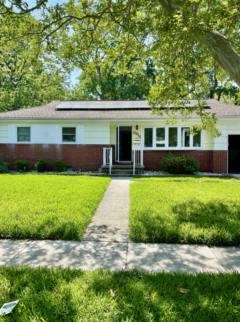 Front view of a house with a brick and yellow exterior, solar panels on the roof, a sidewalk leading to the front door, and lush green lawn and trees.