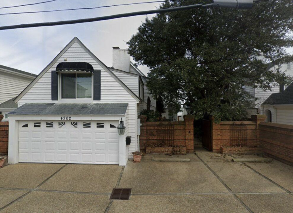 Front view of a white garage house with a black shuttered window, a small lawn area, a brick wall with a garden gate, a large tree, and a driveway.