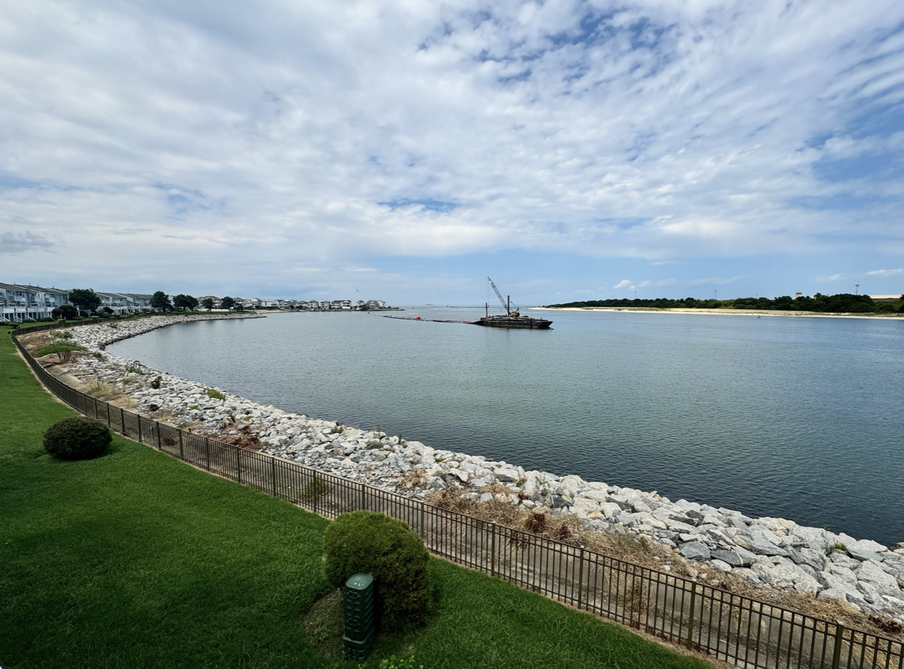 A peaceful waterfront scene with a curved rocky shoreline, green grass, and residential buildings on the left. A barge with a crane is on the water, and there's a partly cloudy sky above.