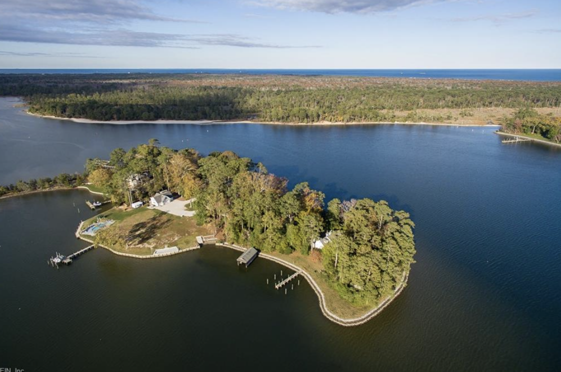 Aerial view of a small peninsula with houses, trees, docks, and boats, surrounded by a large body of water, with a forested area in the background under a partly cloudy sky.