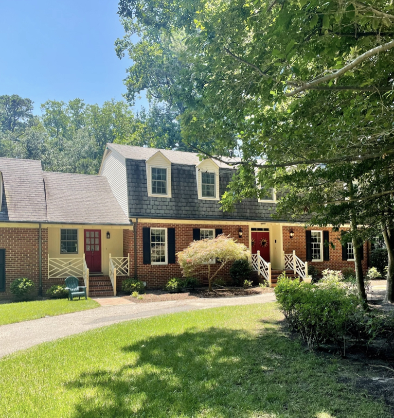 A two-story brick house with multiple gabled windows, a red front door, white porch railings, and surrounded by green trees and a landscaped yard with a lawn and bushes.