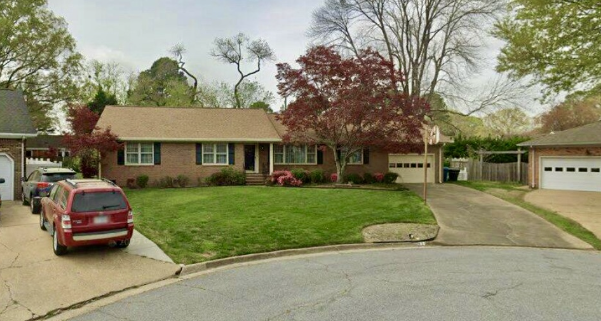 A suburban house with a well-maintained lawn, a driveway with parked cars, and trees in the background.