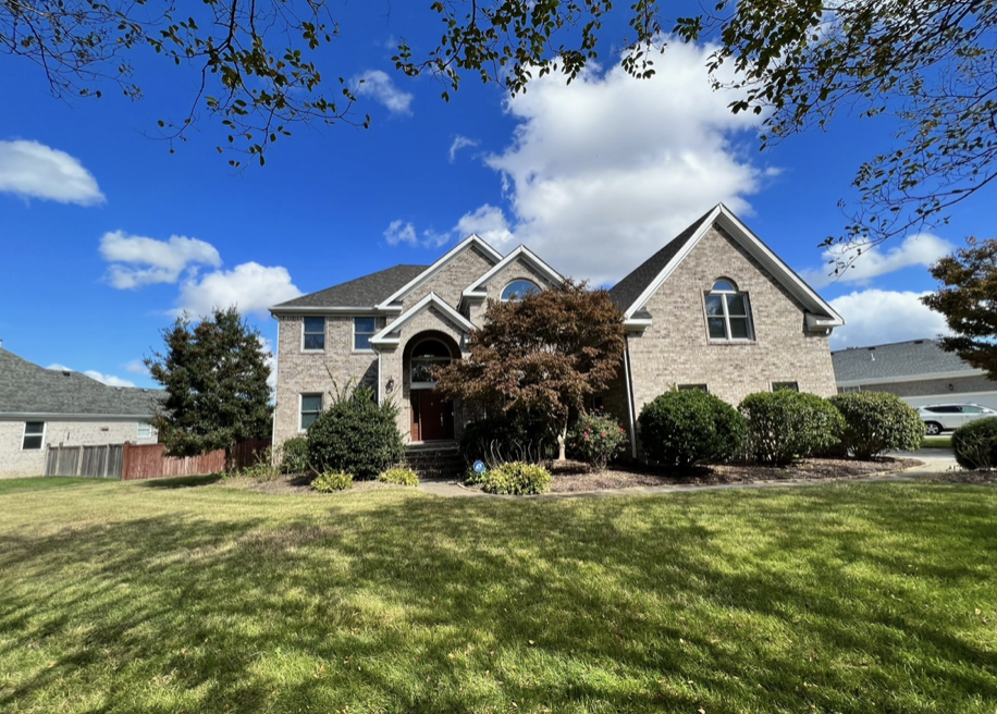 Large brick house with a front porch, surrounded by well-maintained lawn and bushes, under a blue sky with scattered clouds.