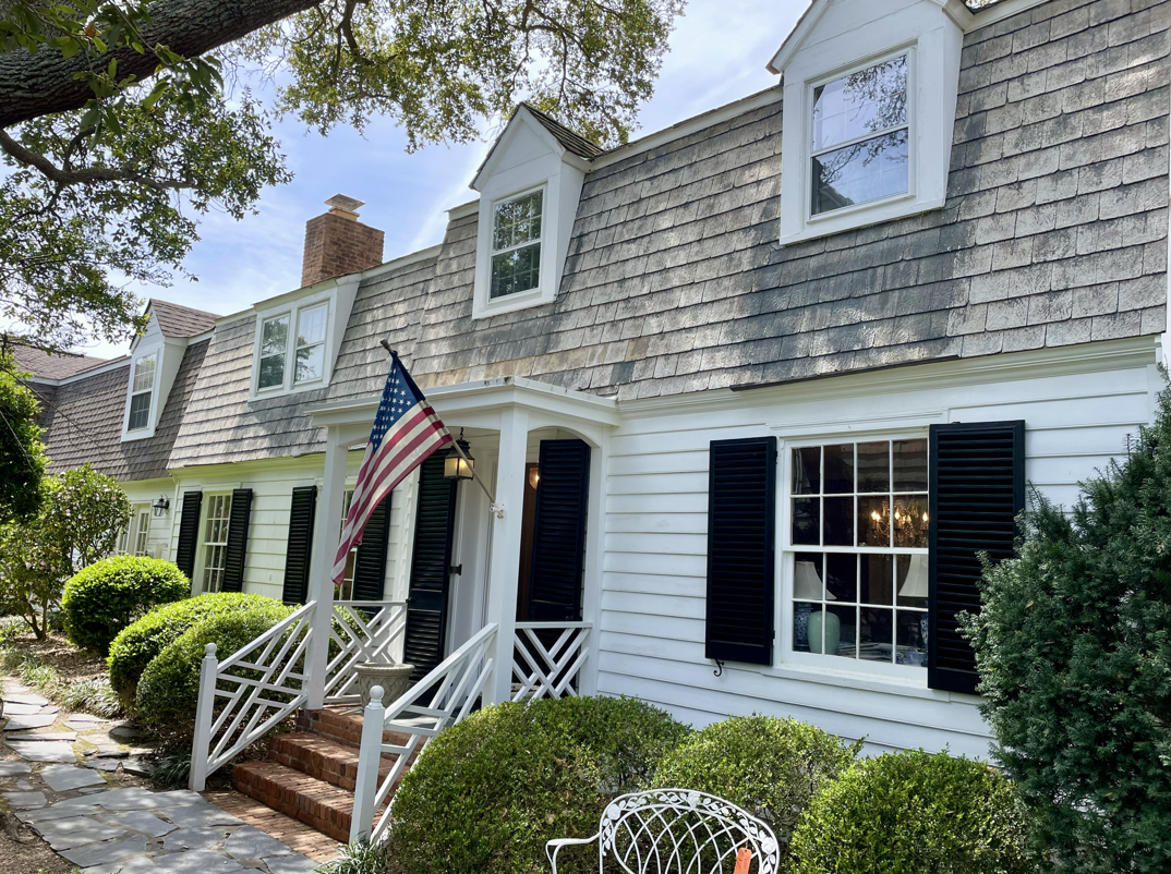White house with black shutters, brick steps, porch light, American flag, lush bushes, trees, and a stone pathway.