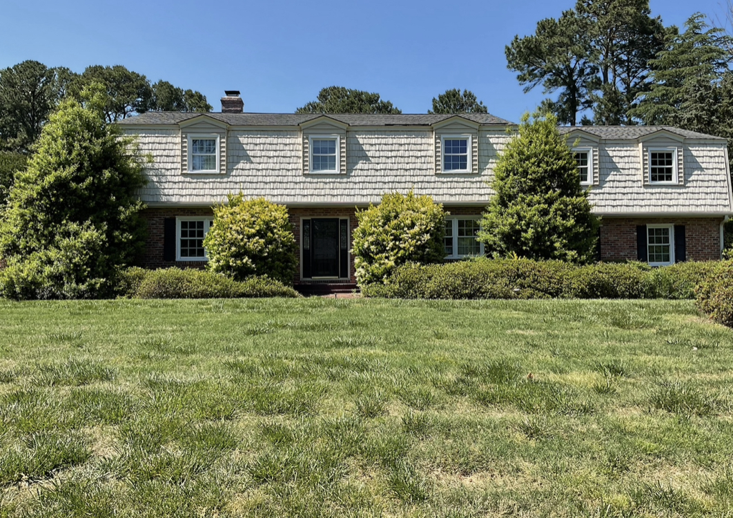 A two-story house with a gray shingle roof, brick exterior, and several windows, surrounded by trees and bushes, with a well-maintained grassy lawn in front.