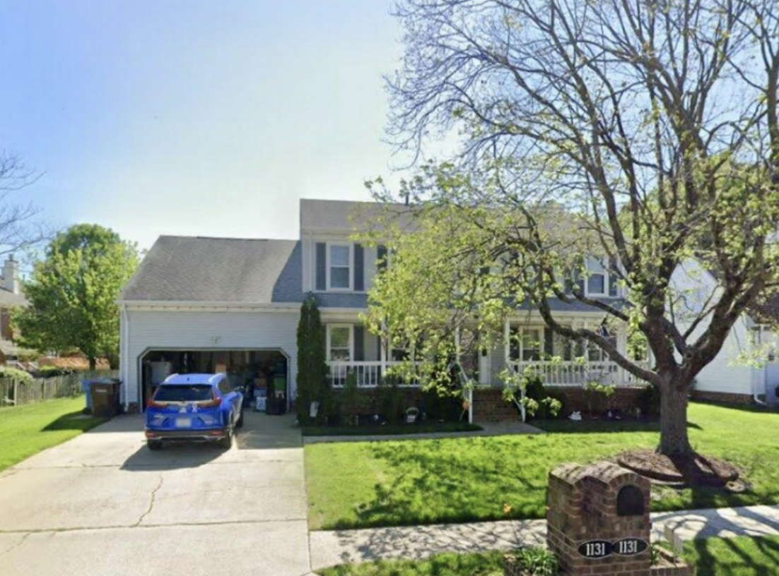 A two-story white house with a garage, a blue car parked in the driveway, and a large tree in the front yard. The house has a porch with white railings, and the front yard is landscaped with a lawn and some bushes.