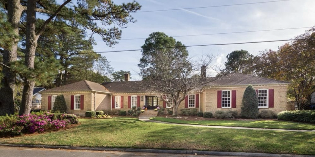 A single-story house with a lawn, trees, and shrubs, featuring beige brick exterior, red shutters, and a pathway leading to the front door.