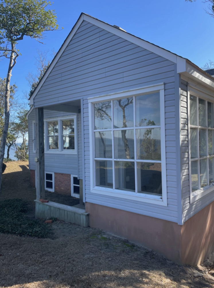 Side view of a house with white vinyl siding, large window, and a small porch. The house is on a grassy lot with trees nearby.