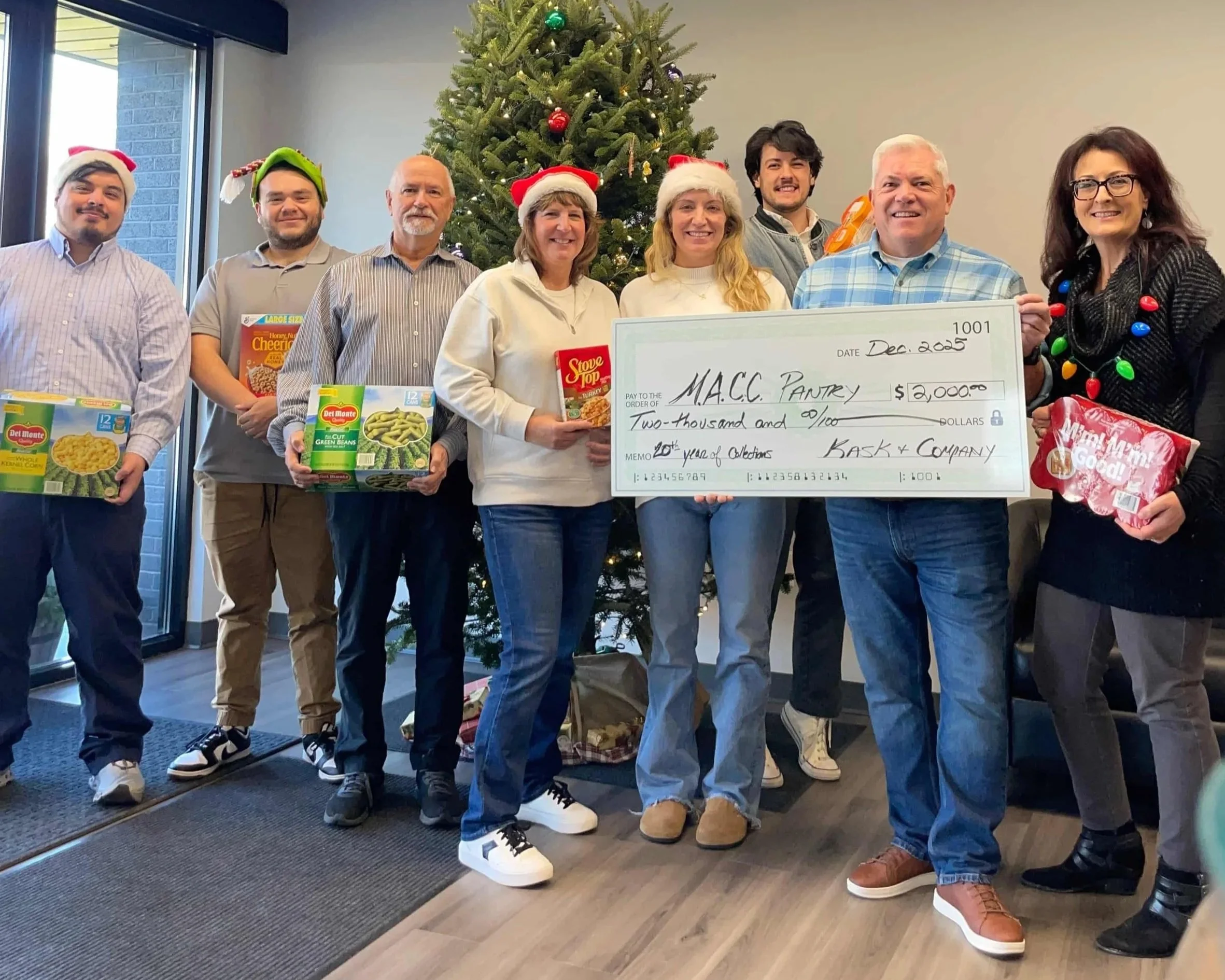 Group of people standing in front of a Christmas tree, holding boxed food items and a large ceremonial check for $2,000 from BASK & COMPANY made out to M.A.C.C. Pantry, decorated with holiday hats and casual clothing, inside a room with a window and a decorated tree.