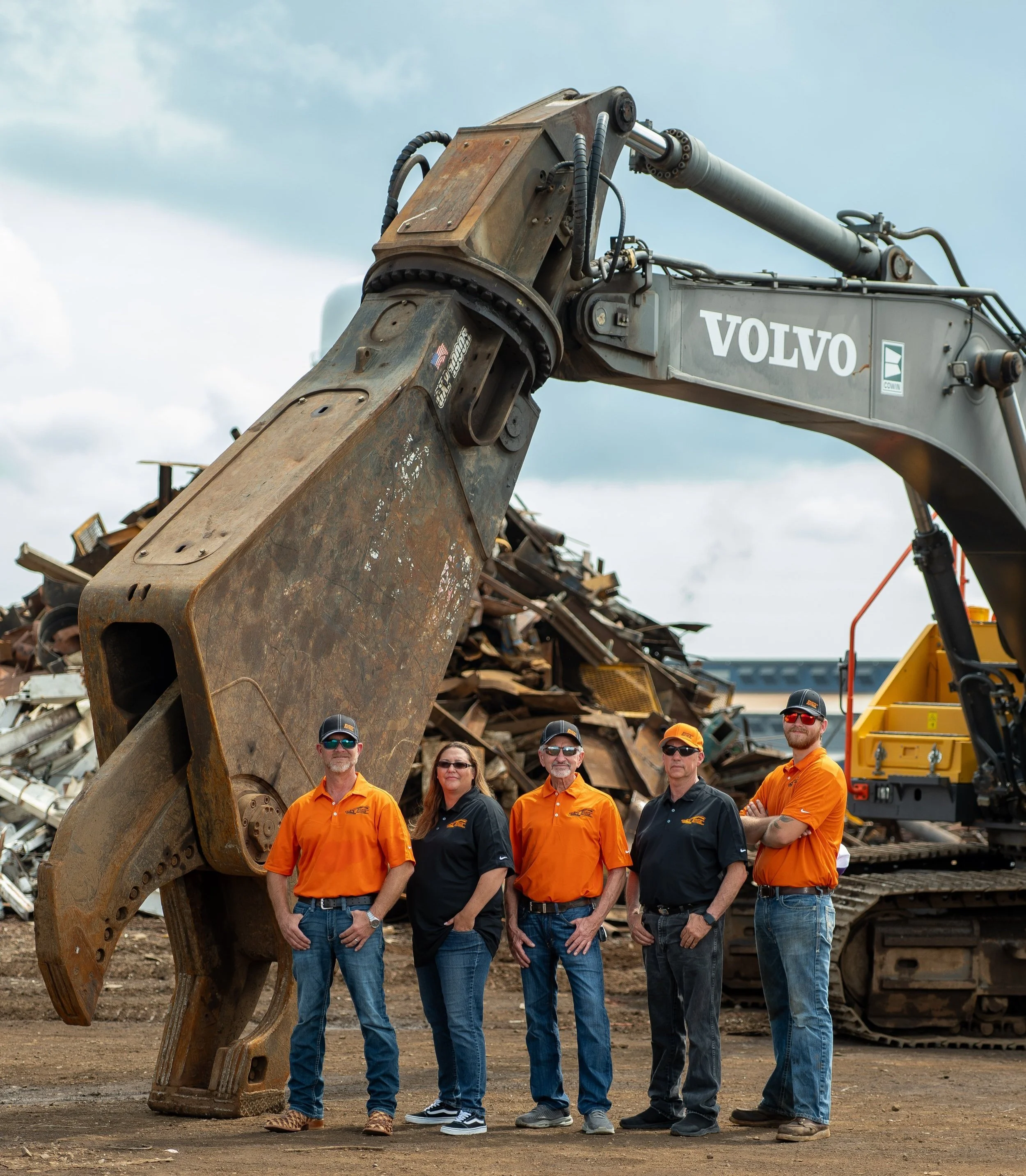 Five people standing in front of a large excavator and a pile of scrap metal, wearing safety glasses and work shirts, at an industrial site.