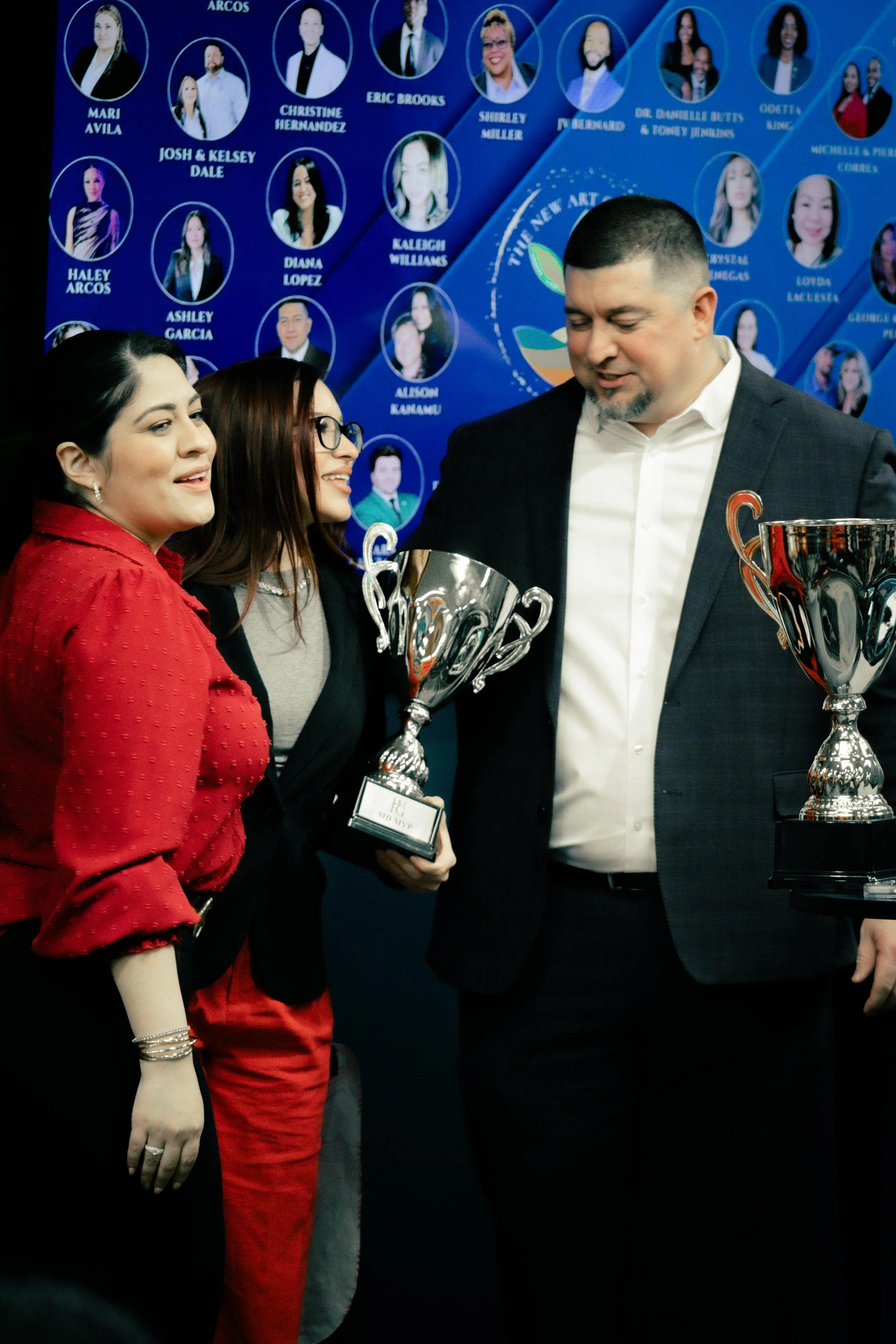 Three people, two women and one man, are standing together, smiling, and holding trophies. They are at an awards or recognition event, with a backdrop displaying photographs and names of other individuals.