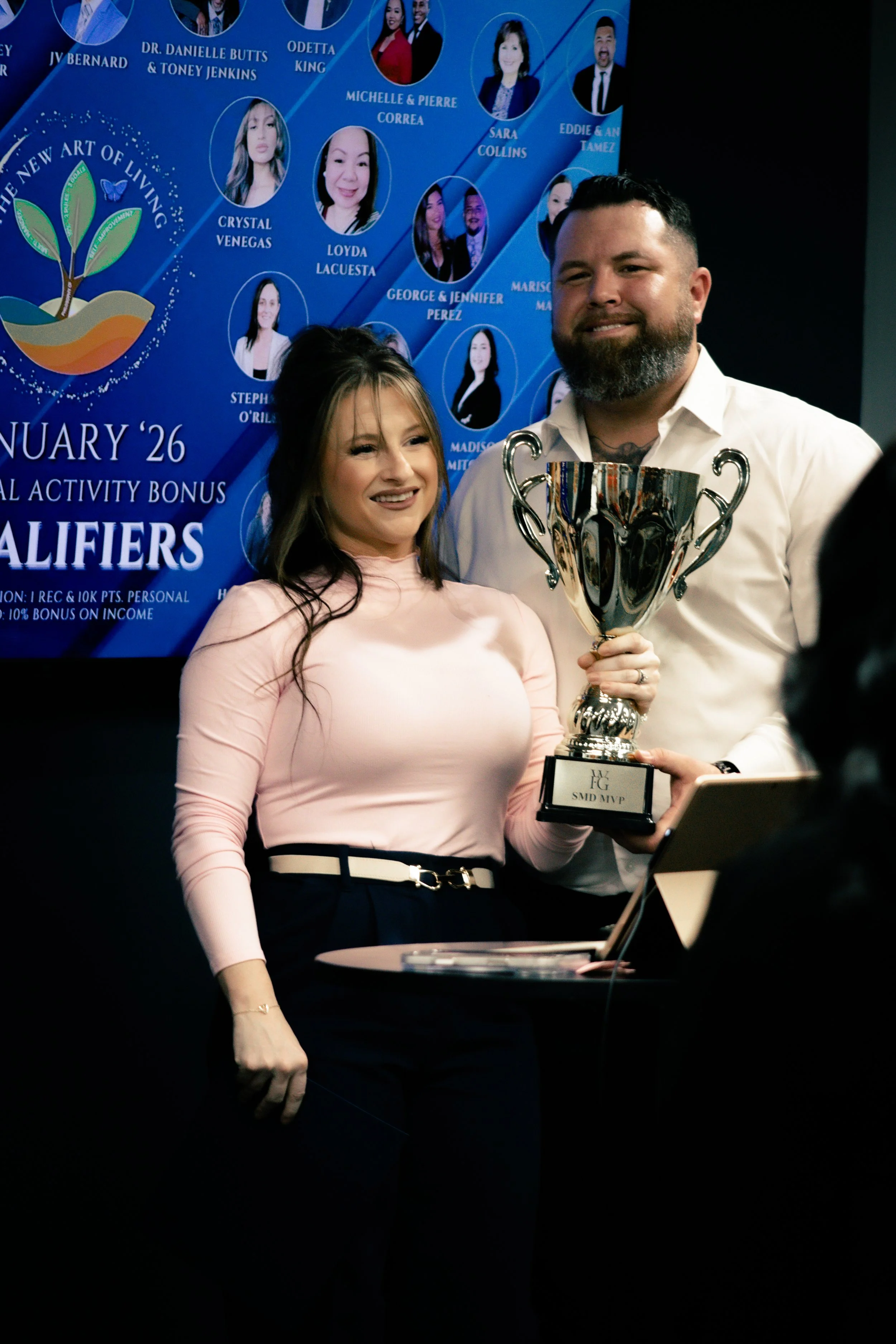 A woman and a man smiling as they hold a large trophy together during an awards ceremony, with a blue poster showing photos and names of people in the background.