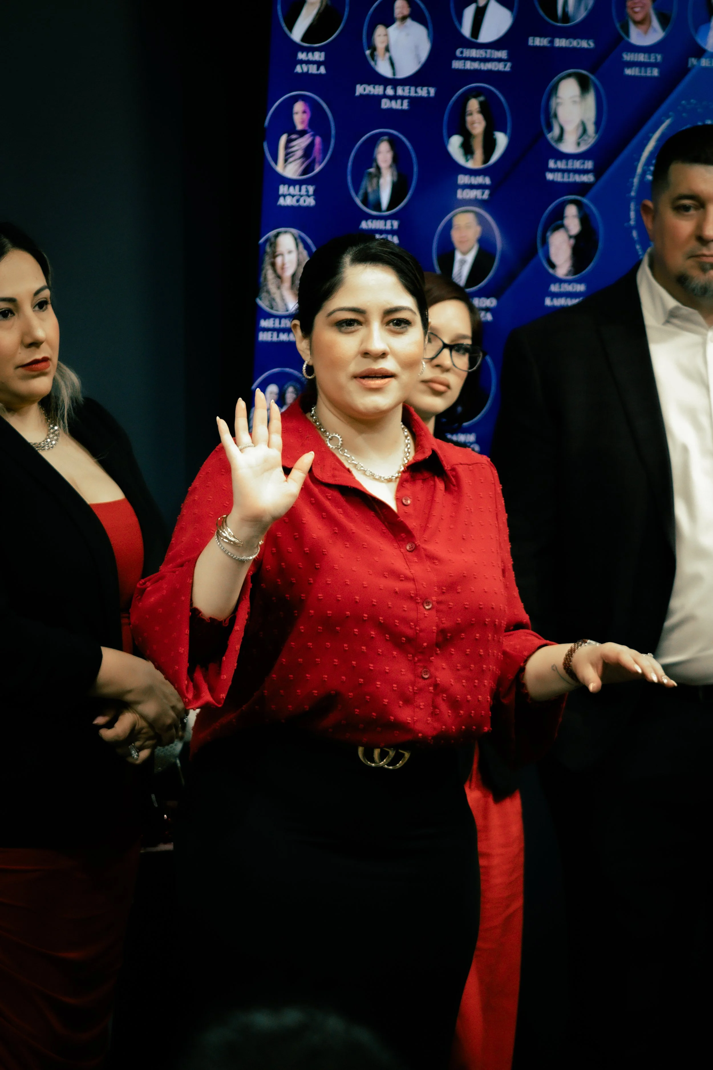 Woman in red blouse with jewelry raising her right hand at a formal event. Other people stand around her, with a background featuring a blue display with headshots and names.