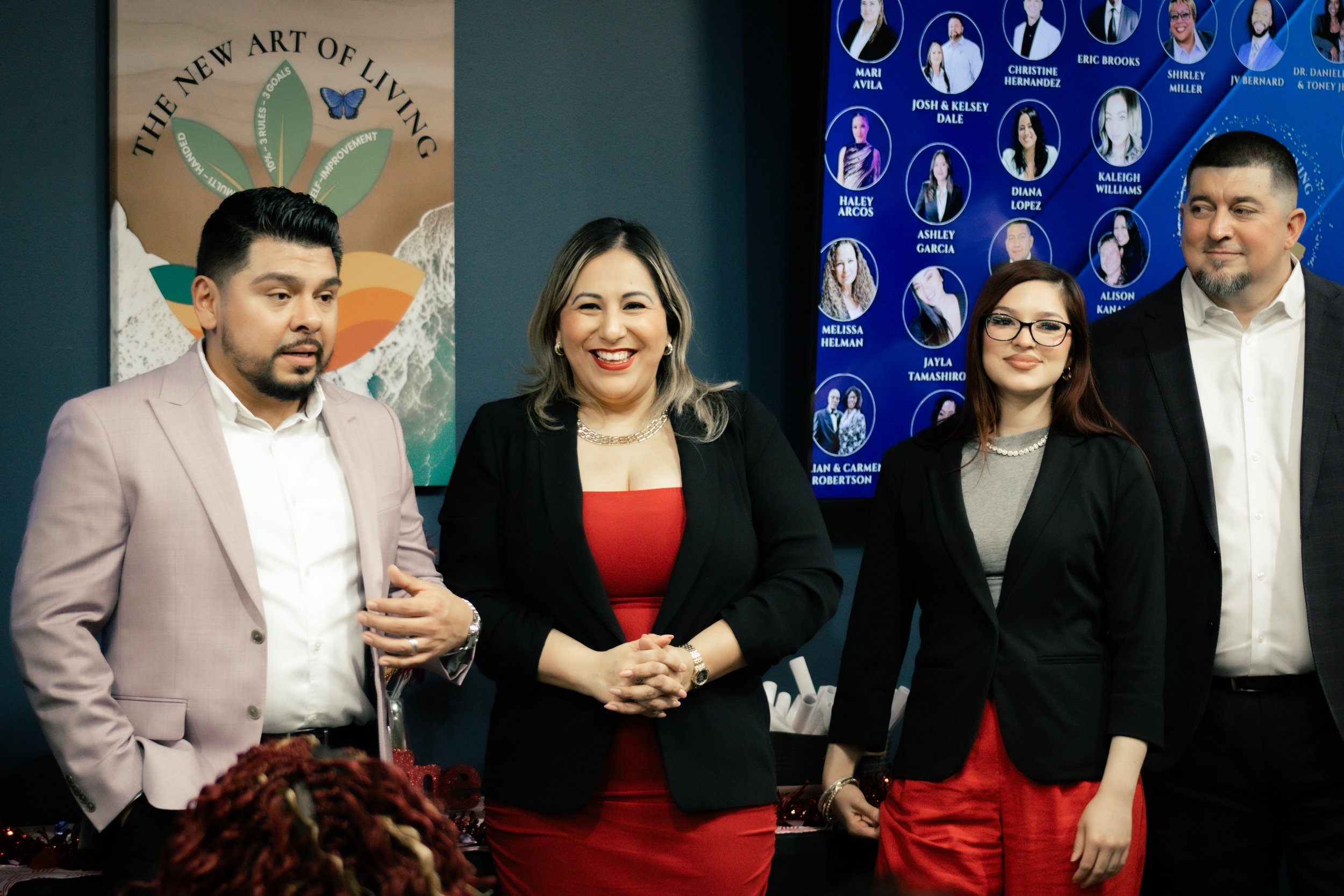 Group of four people standing indoors in front of posters, wearing business attire, smiling and posing for the photo.