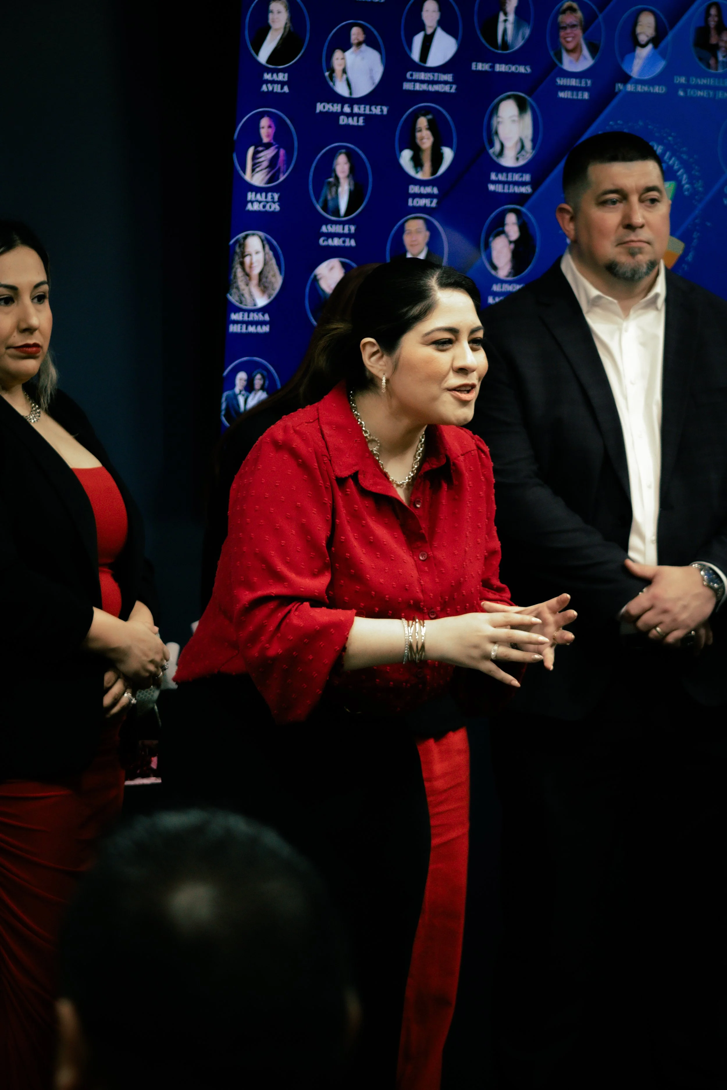 A woman in a red blouse speaking, standing next to a man in a suit and a woman in a black blazer. There is a blue poster with photos of other people behind them.