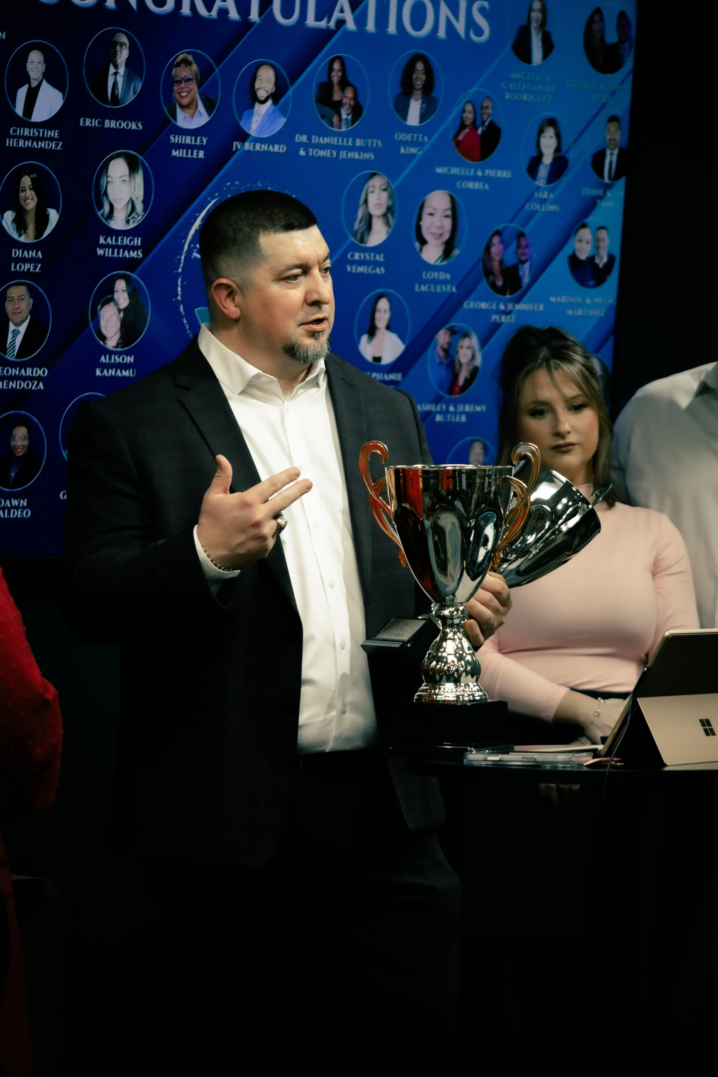 A man in a suit holding a trophy and a woman in a pink dress standing behind a table with a laptop. There is a large blue congratulatory poster with photos of multiple people in the background.