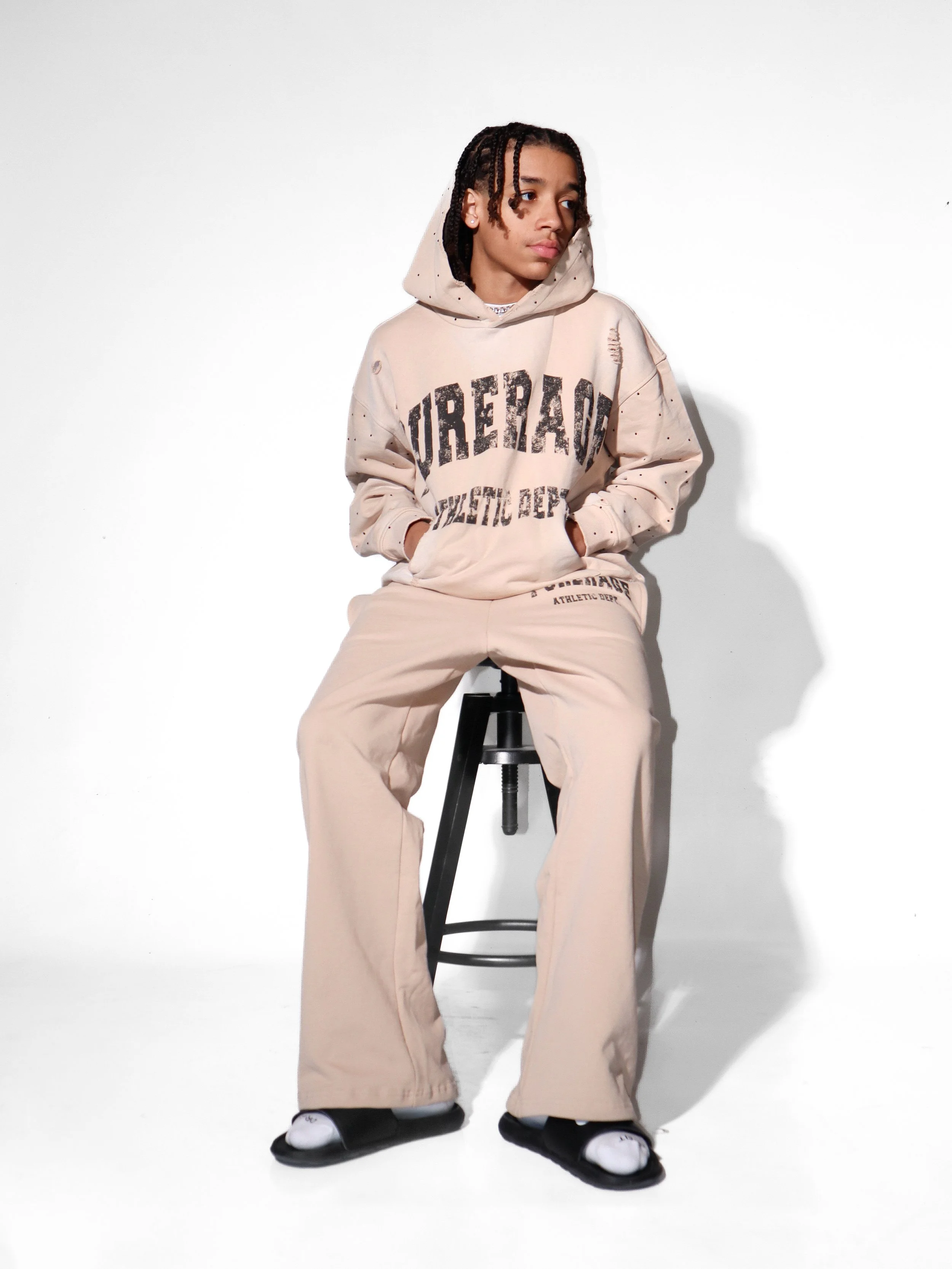 A person sitting on a black stool against a white background, wearing a beige hoodie with black text and beige wide-leg pants. They have their hands in their hoodie pockets and are wearing slide sandals over white socks.