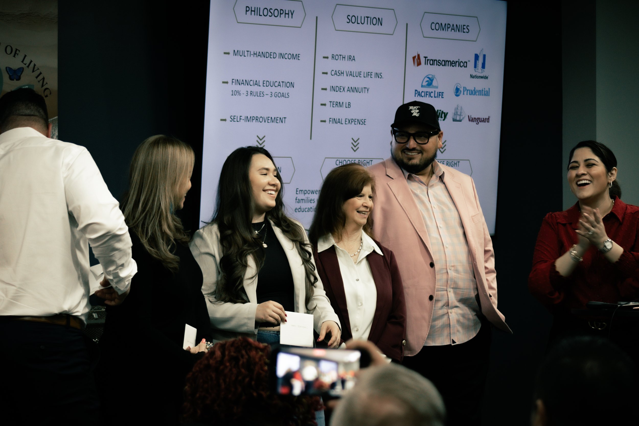 A group of smiling people standing in front of a presentation screen, with one woman clapping. The presentation screen behind them displays categories such as 'Philosophy,' 'Solution,' and 'Companies' with various bullet points and company logos.