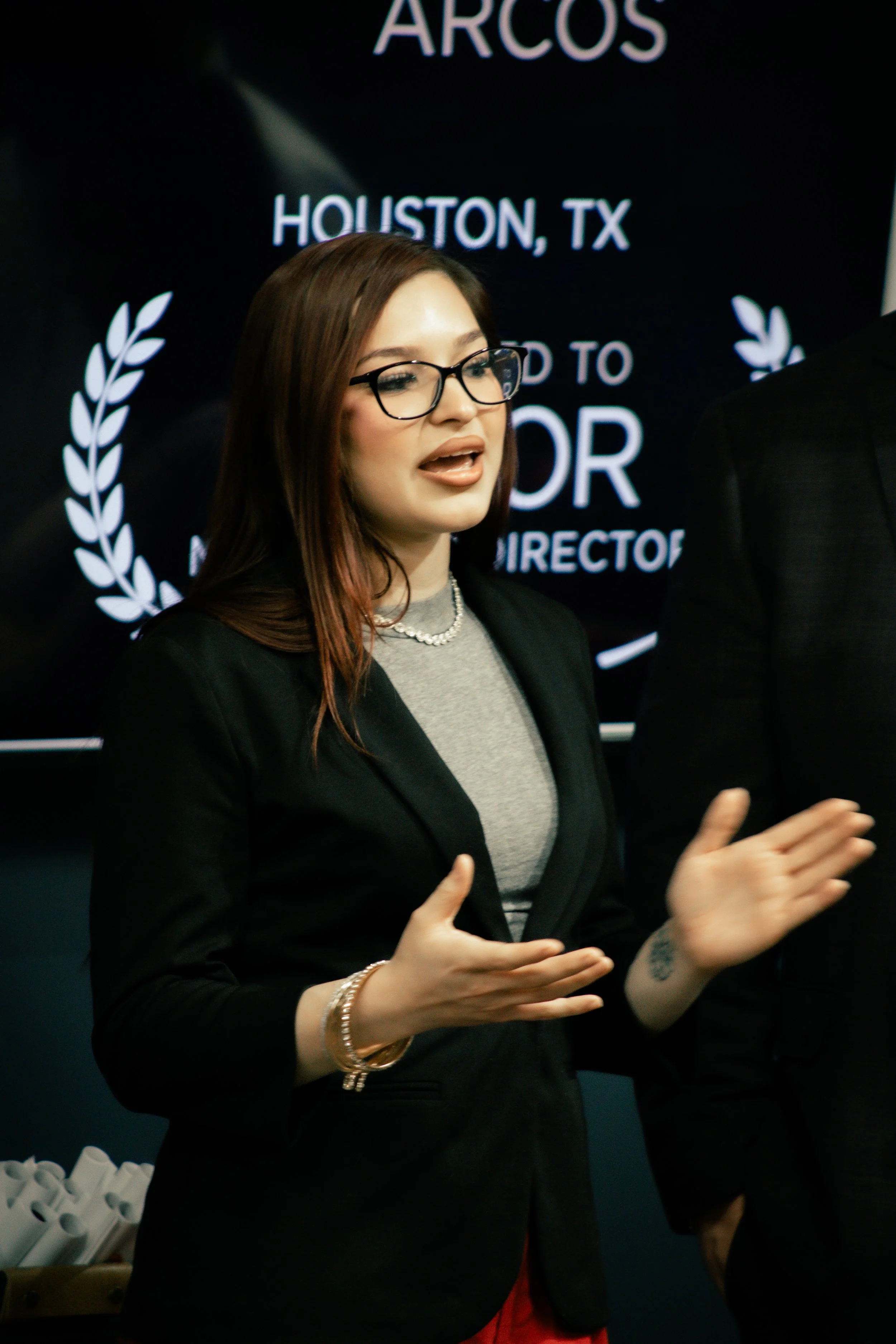 A woman with glasses and long brown hair speaking at an event, with a black backdrop featuring white text and laurel designs.