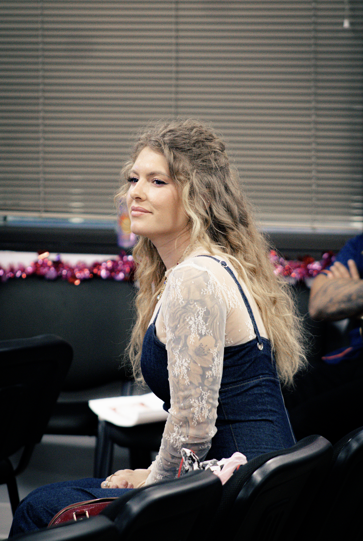 A young woman with long, wavy blonde hair sits on a black seat in a room decorated for Christmas, with garland and ornaments visible in the background.