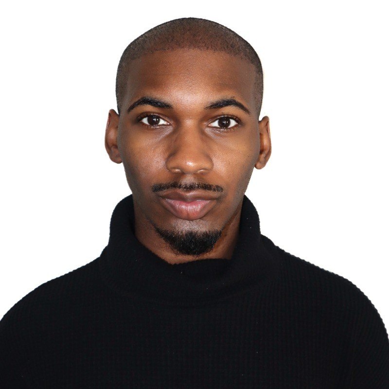 A young man with dark skin, short hair, and a goatee, wearing a black turtleneck sweater, looking directly at the camera with a neutral expression.