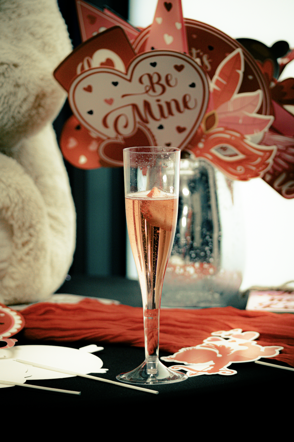 A glass of sparkling rosé champagne with a slice of strawberry inside on a table with Valentine's Day decorations, including a heart-shaped sign that says 'Be Mine' and other Valentine's themed items in the background.