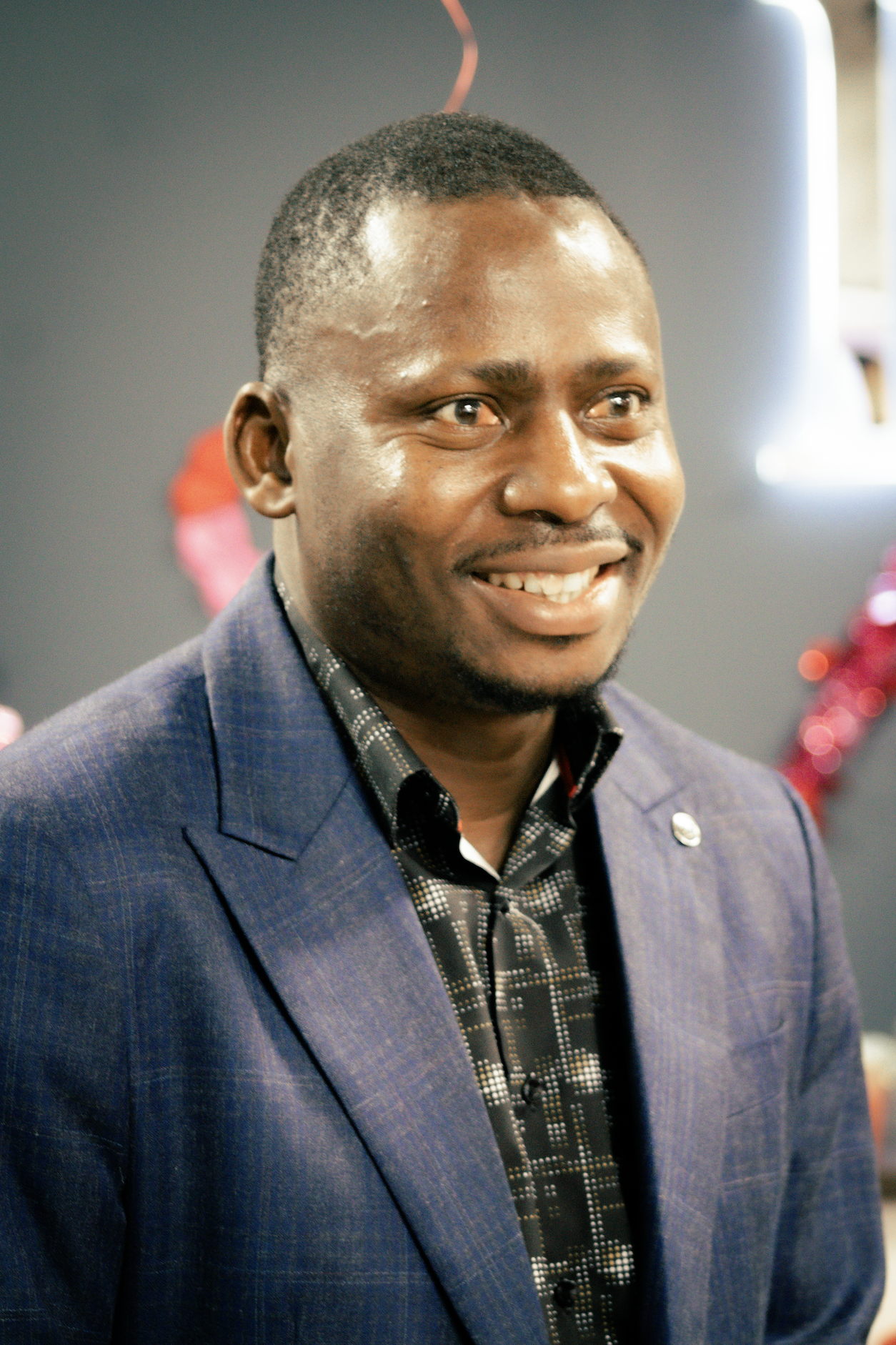 A smiling man in a blue blazer and patterned shirt at an indoor event.