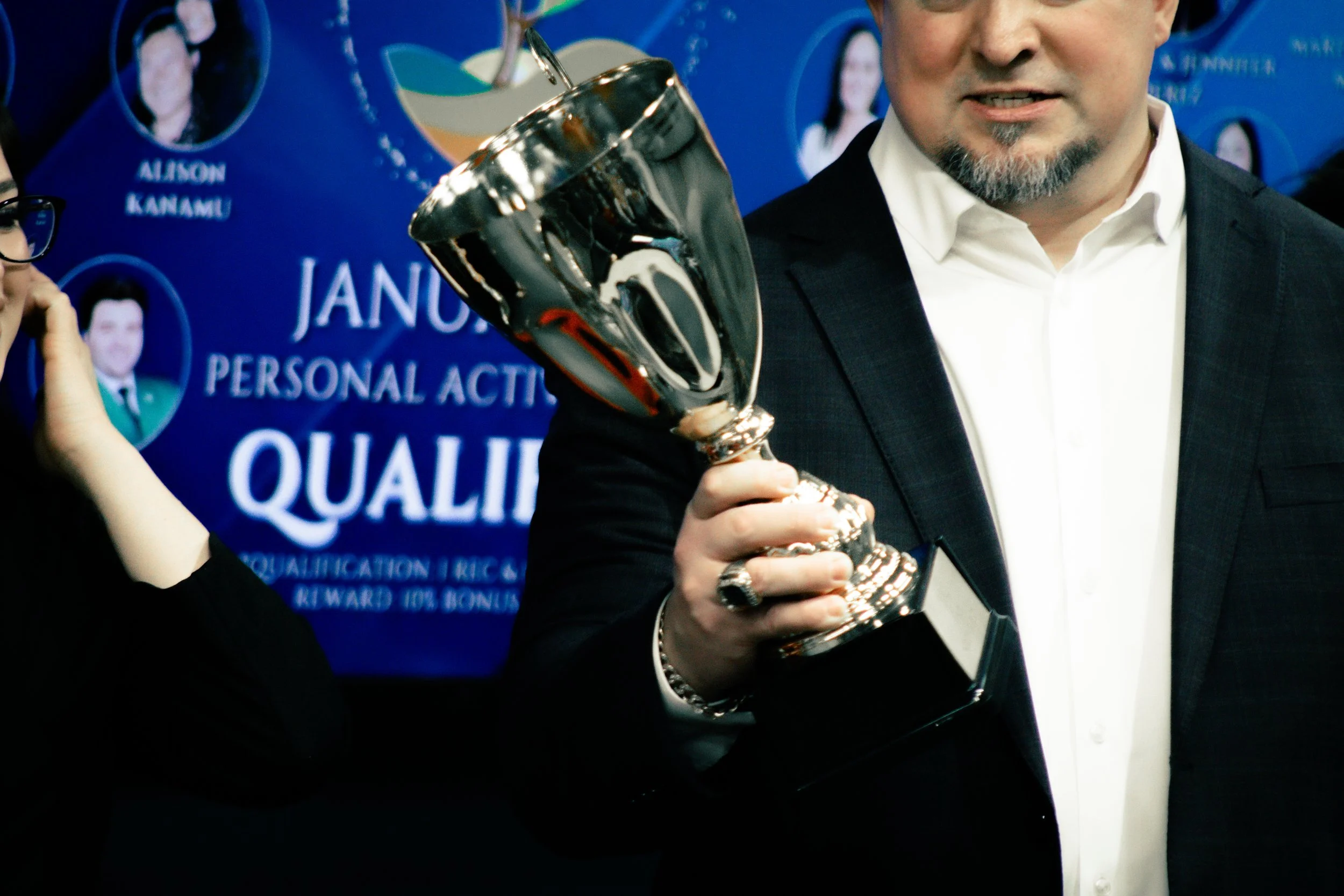 A man in a white shirt and dark suit holding a shiny silver trophy at an awards event with a blue background.