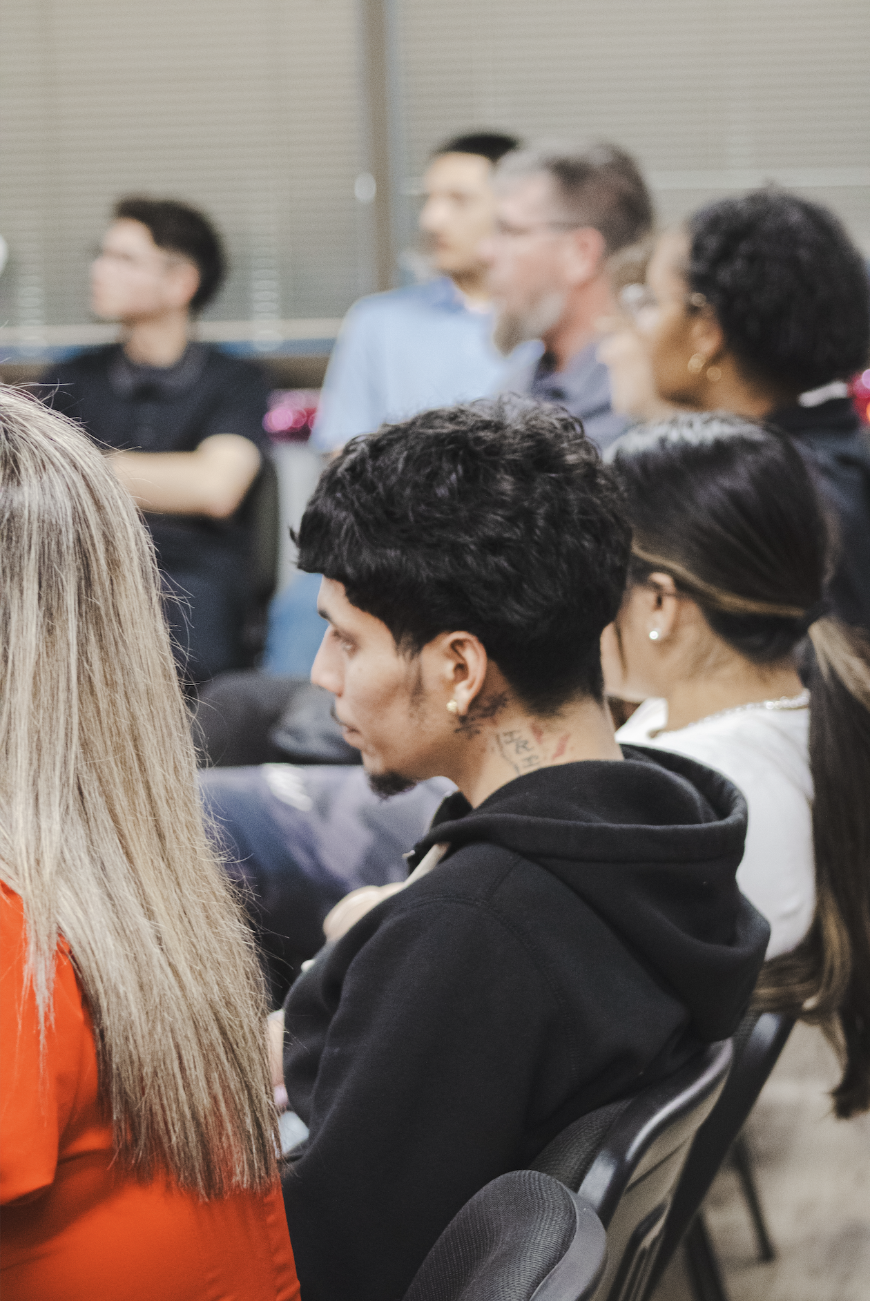 Audience seated, listening attentively at an indoor event, with diverse individuals including a man with curly black hair and tattoos on his neck in the foreground.