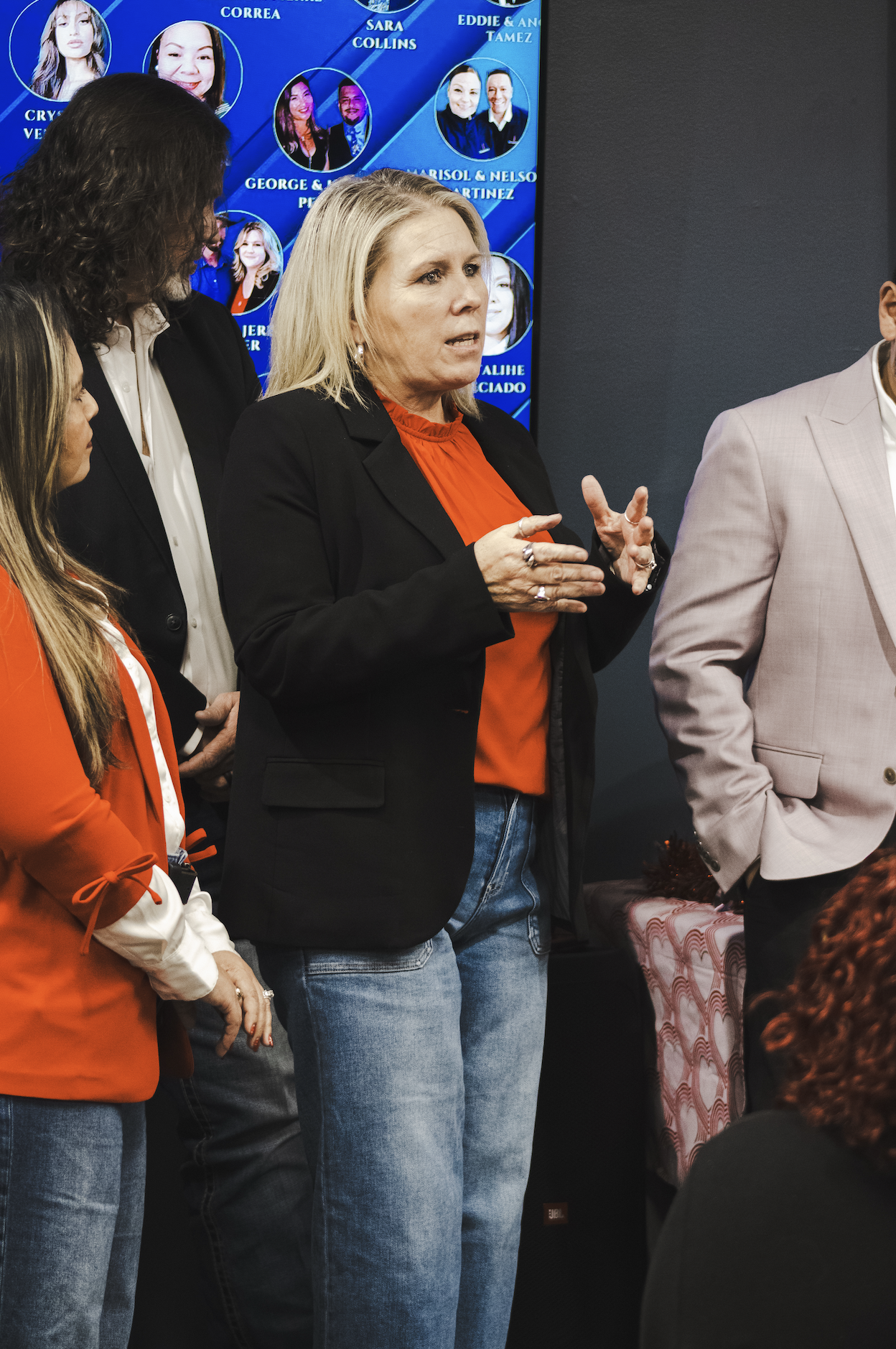 Woman speaking at an indoor event, wearing a black blazer and orange top, surrounded by other people, with a blue background displaying headshots and names.